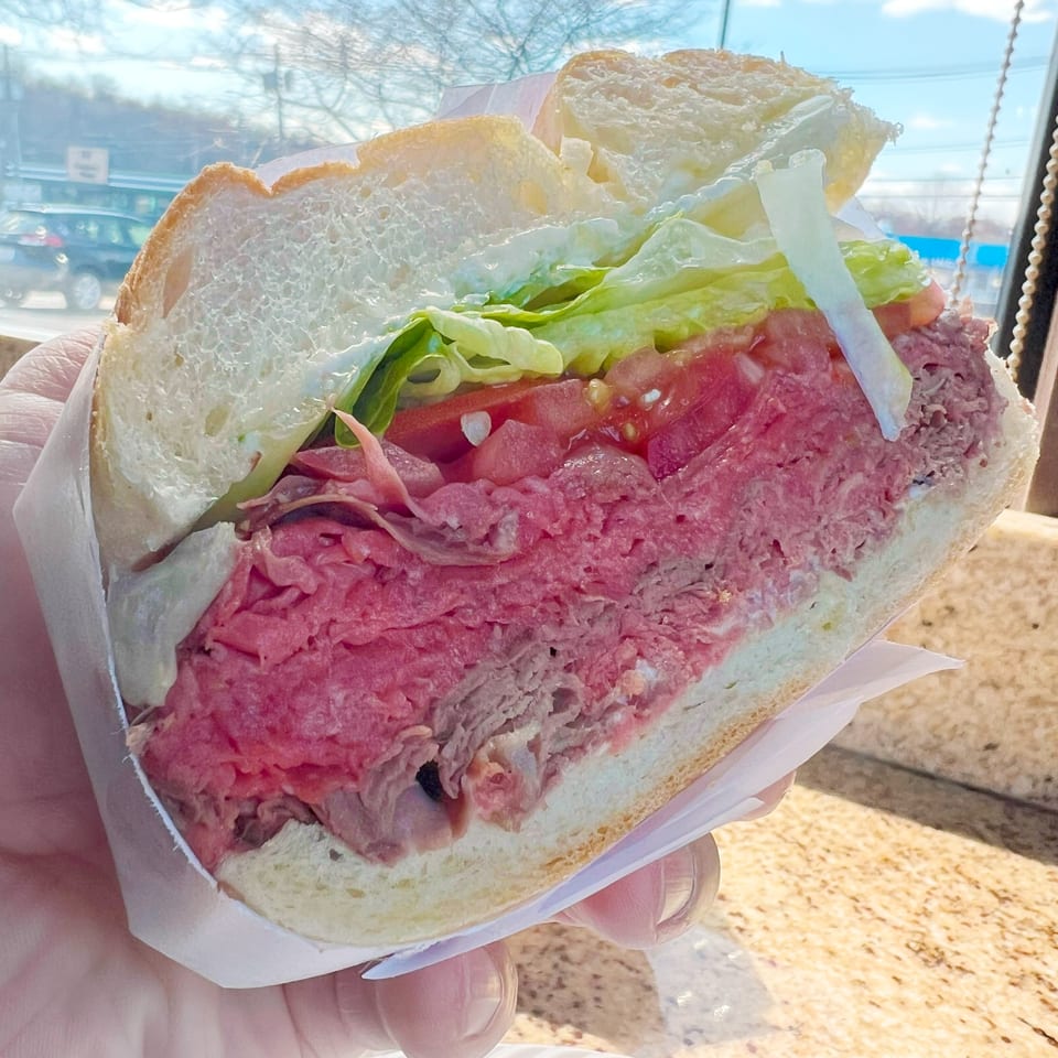 A hand holds a large Bagel Boys roast beef sandwich with lettuce and tomato, outdoors.