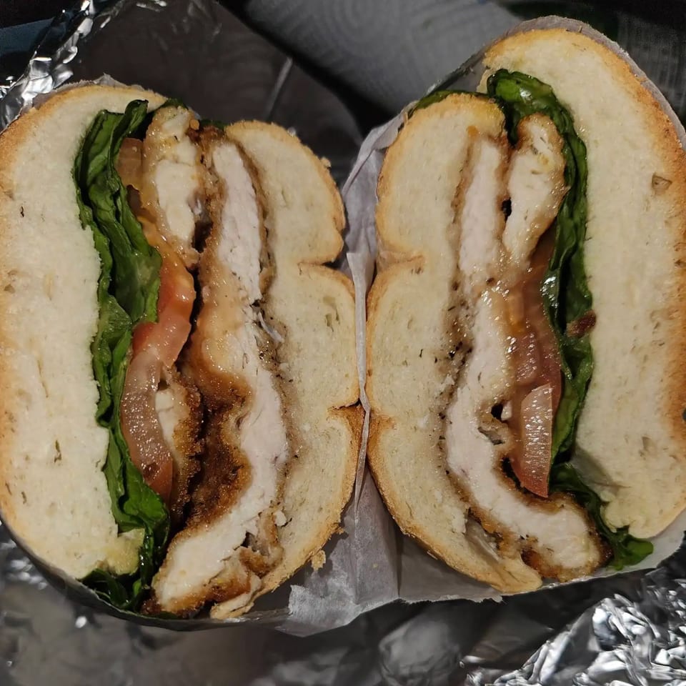 A Bagel Boys bagel sandwich, cut in half, revealing crispy chicken, lettuce, tomato, and sauce.